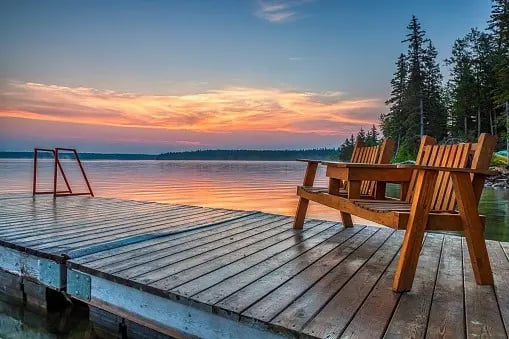 Wooden lake dock with two Adirondack chairs facing calm water at sunset.