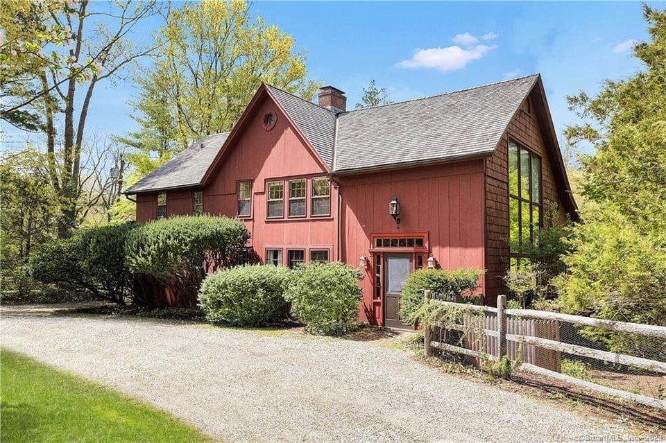 Red two-story house with a gravel driveway, clipped shrubs, and a low wooden fence.