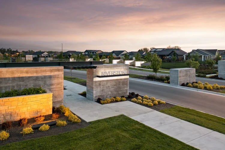 Riverstone neighborhood entrance sign beside a road, with stone walls and homes under a pink dusk sky.