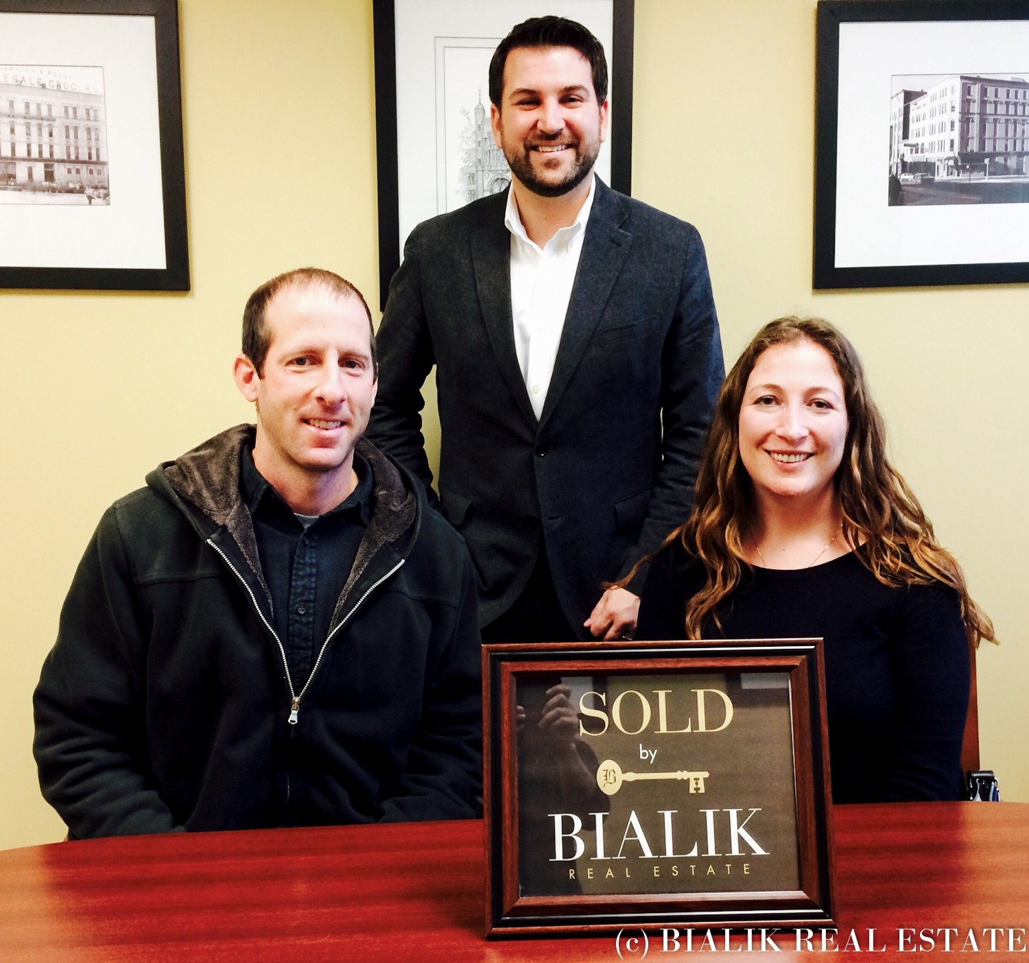 Keith and Cara pose with their real estate agent beside a framed SOLD sign.
