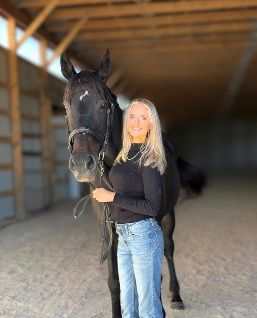 Marie Halbach standing beside a bridled black horse inside a stable.