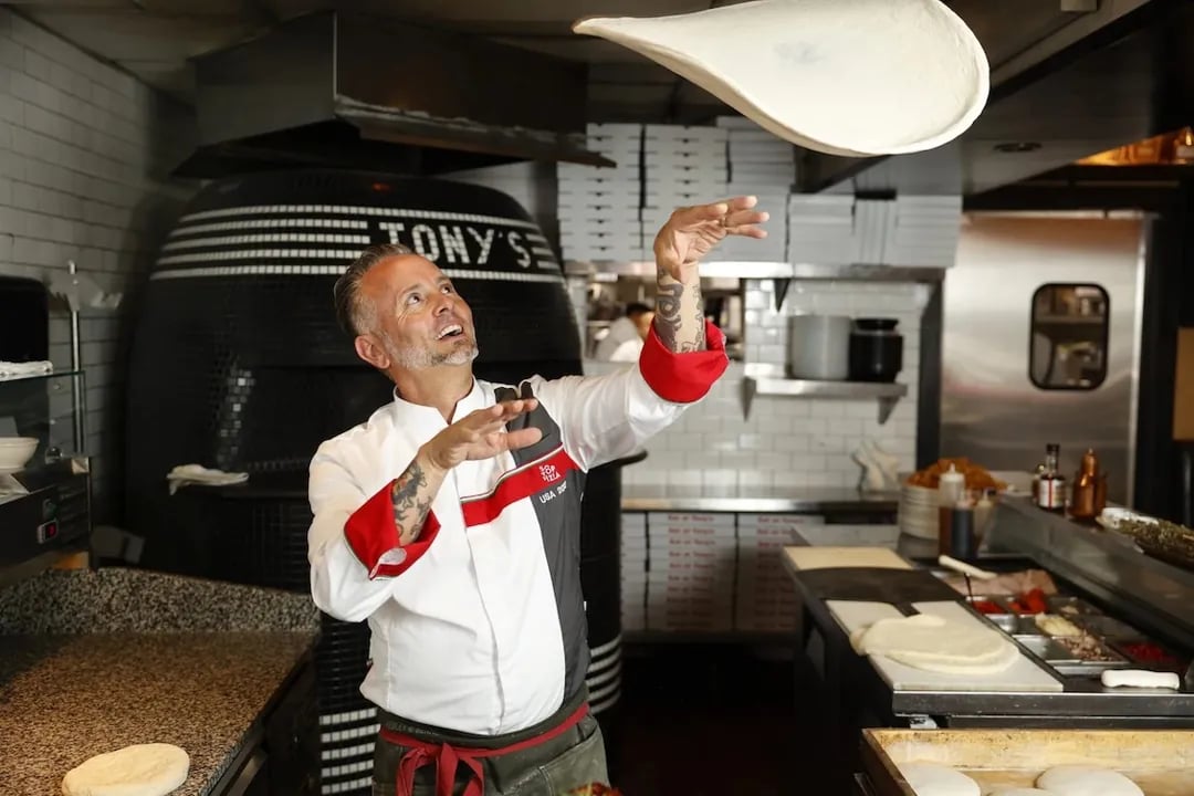 Close-up of hands tossing pizza dough in the air with Tony Gemignani demonstrating pizza-making techniques to an engaged crowd