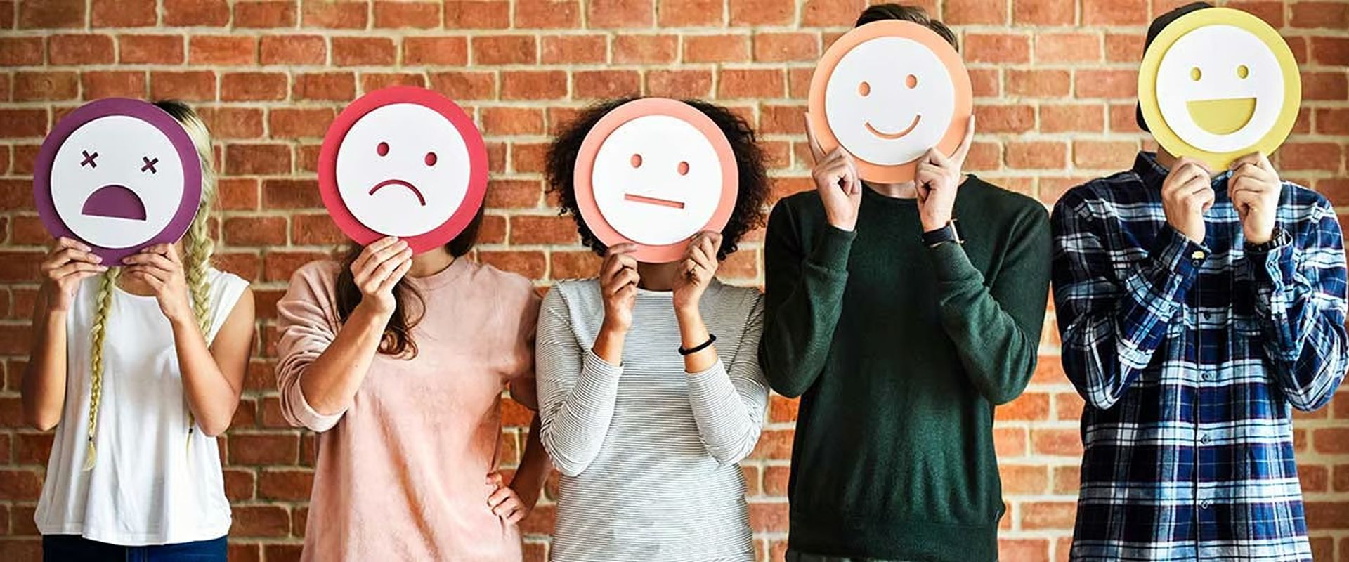 Five people hold expression signs ranging from distressed to happy against a brick wall.