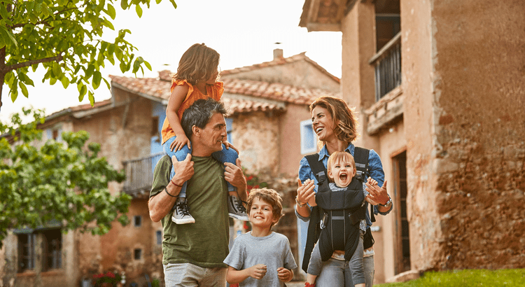 A joyful family strolls outside, with a child on the dad's shoulders and a toddler in a mom's carrier. They smile warmly, surrounded by rustic homes and greenery.