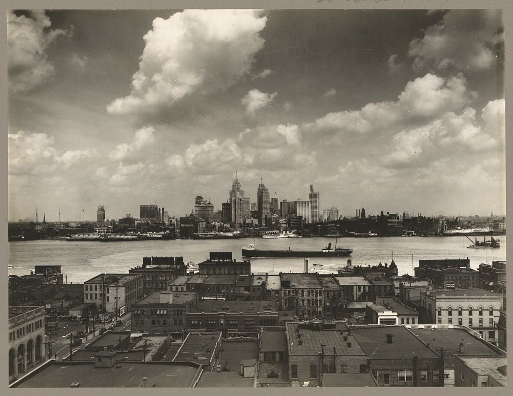 Historic Detroit skyline across the river, with ships on the water and waterfront buildings in the foreground.