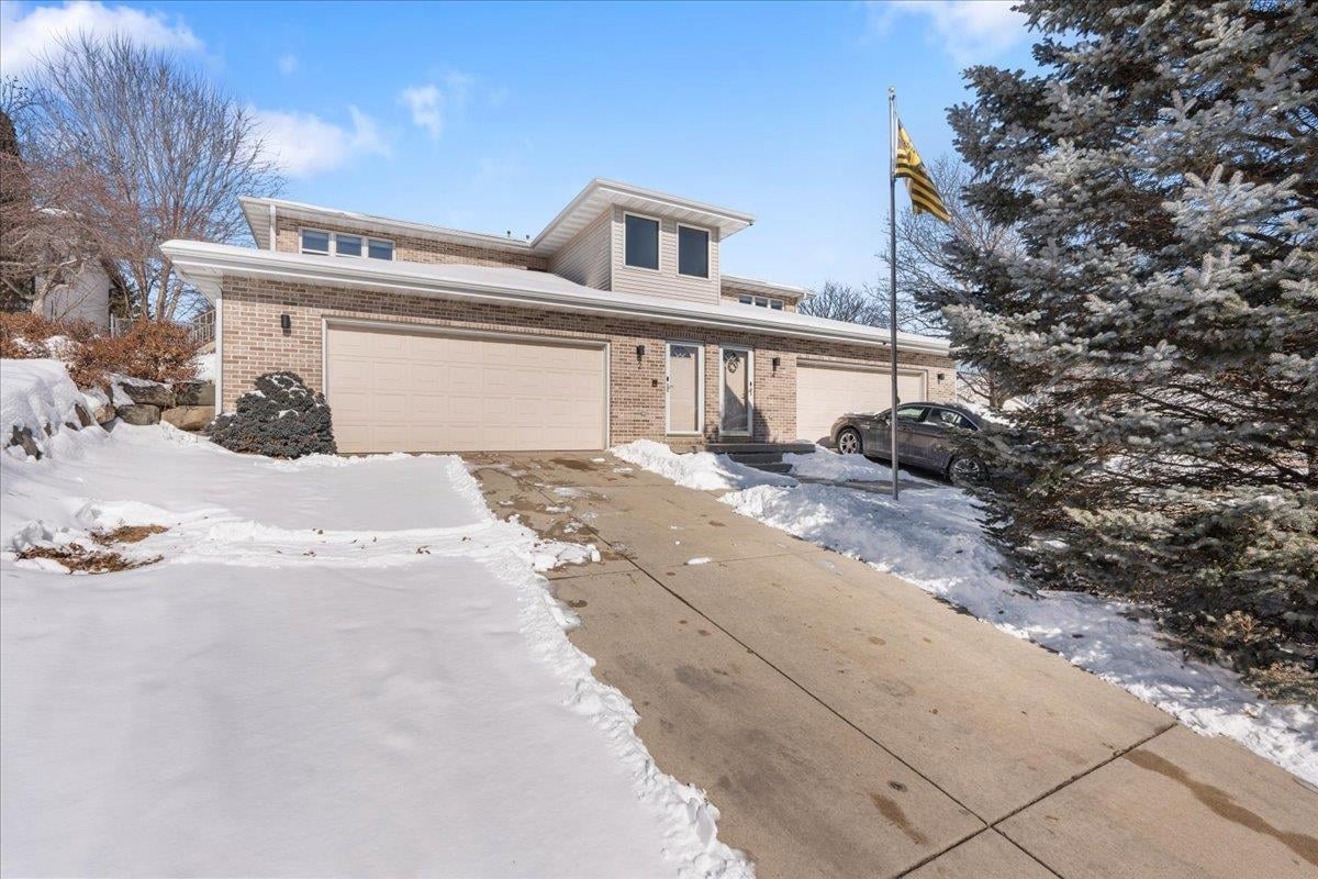 A brick two-story Madison, WI home in winter with snow on the roof and eaves, snowbanks along a cleared driveway, and a frosted evergreen.