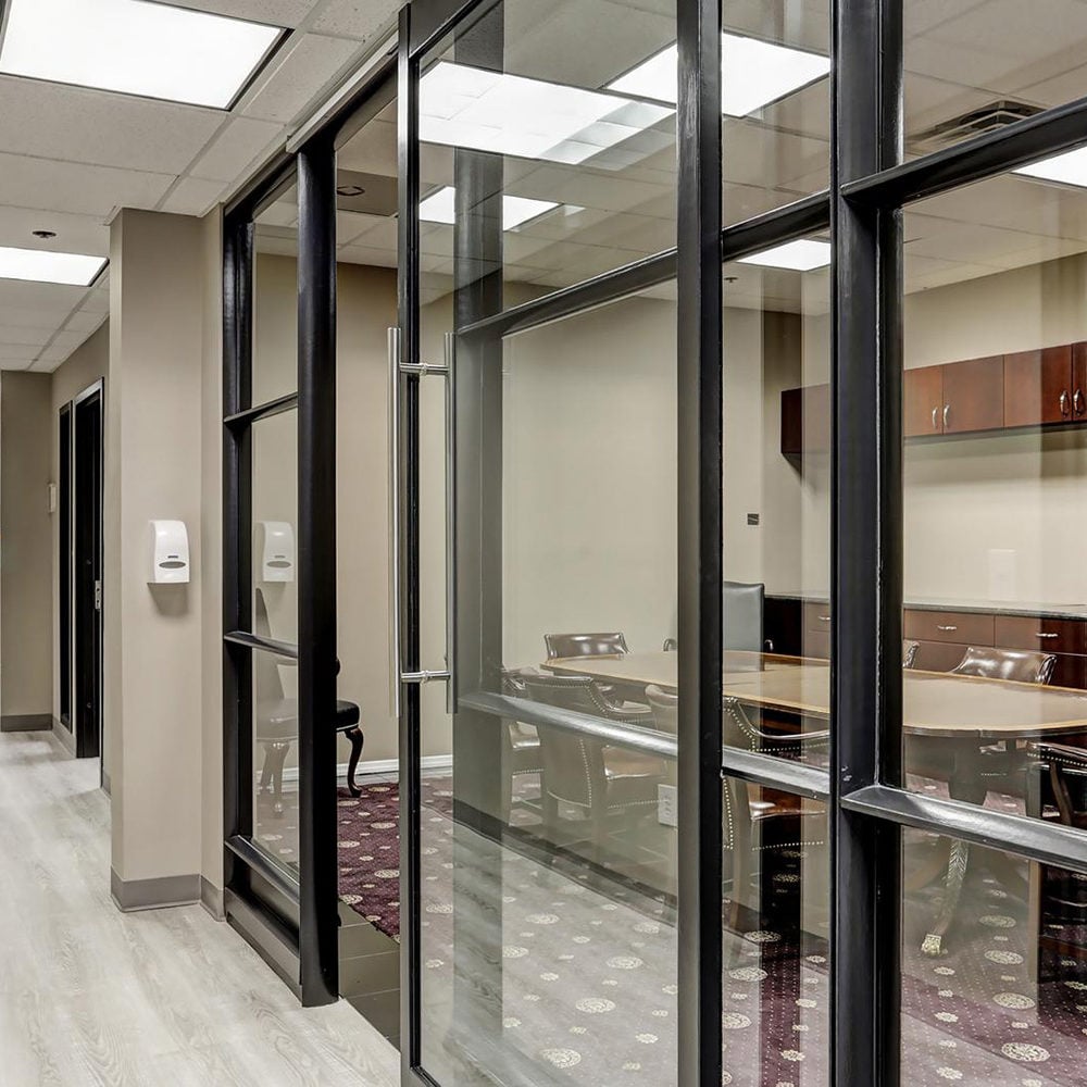 Office conference room behind black-framed glass, with a conference table and wood cabinets.