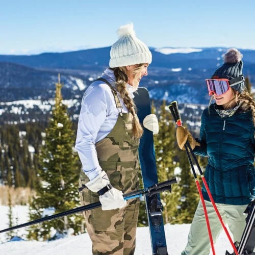 Two women in ski gear with their skis on a snowy mountain slope.