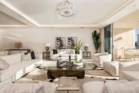 Bright staged living room with white sectional sofas, glass coffee table, and balcony doors.