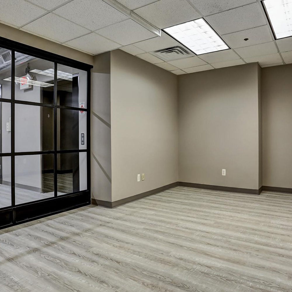 Vacant interior with wood-look flooring and a black-framed glass partition beside an elevator.