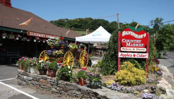 Silverman's Farm Market entrance with flowers, wagon wheels, and a roadside sign for apples and cider.