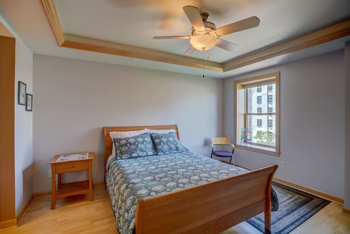 Bedroom with a wooden bed, blue patterned bedding, and a window overlooking a neighboring building.