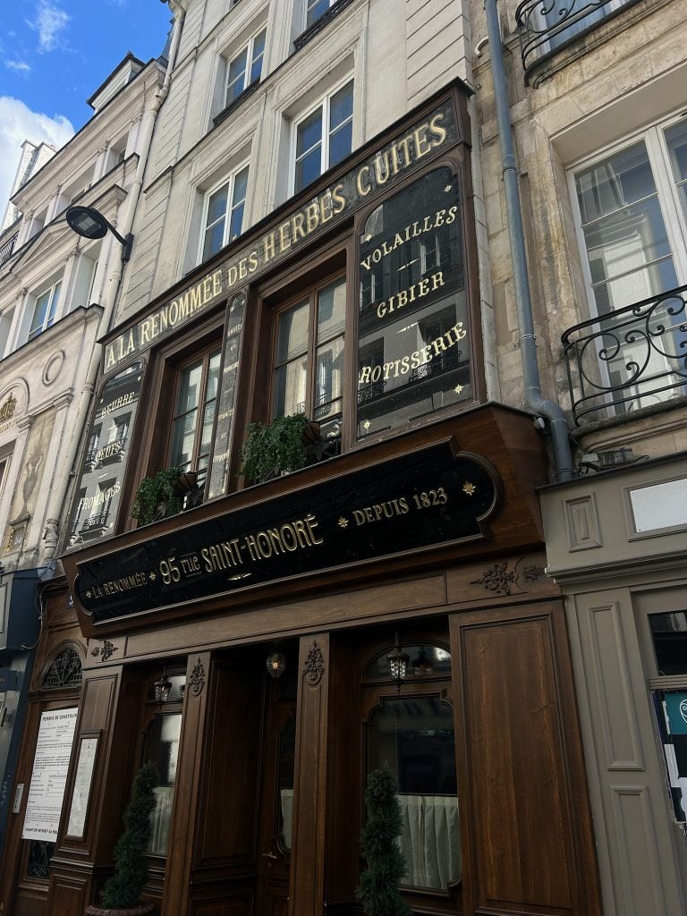 Historic Paris storefront with ornate dark woodwork, gold lettering, and wrought-iron balconies above.