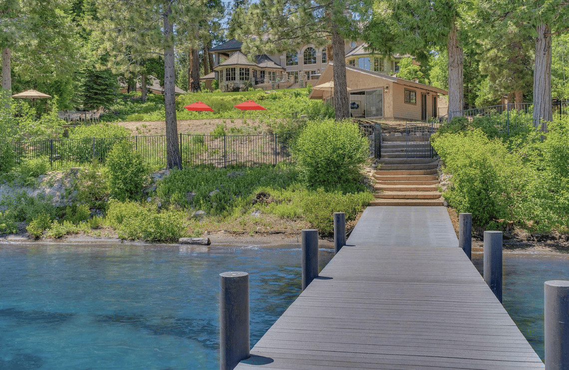 Lakefront home among pine trees, viewed from a dock extending over clear blue water.