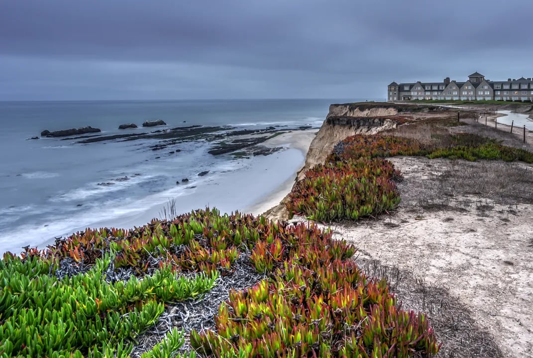 Coastal bluffs with colorful ice plant vegetation overlooking sandy beach and rocky shore, gray hotel building visible on clifftop under overcast sky