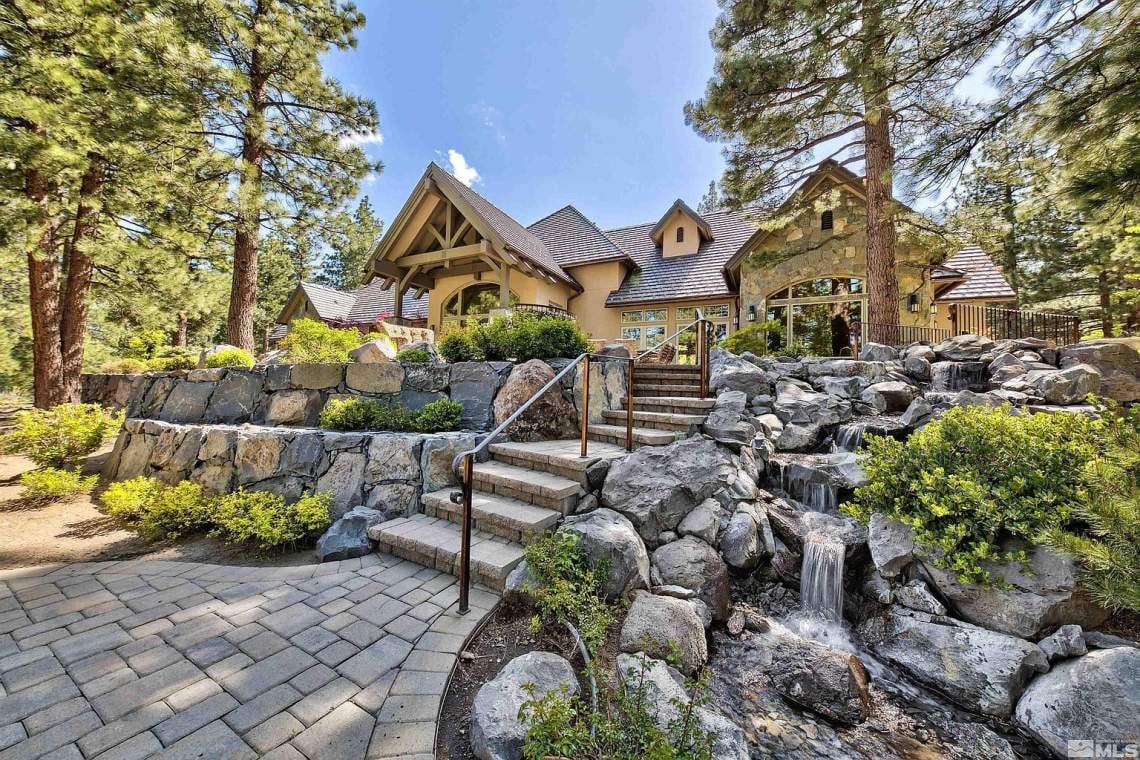 Large mountain-style home framed by pines, with stone entry steps and a landscaped rock waterfall.