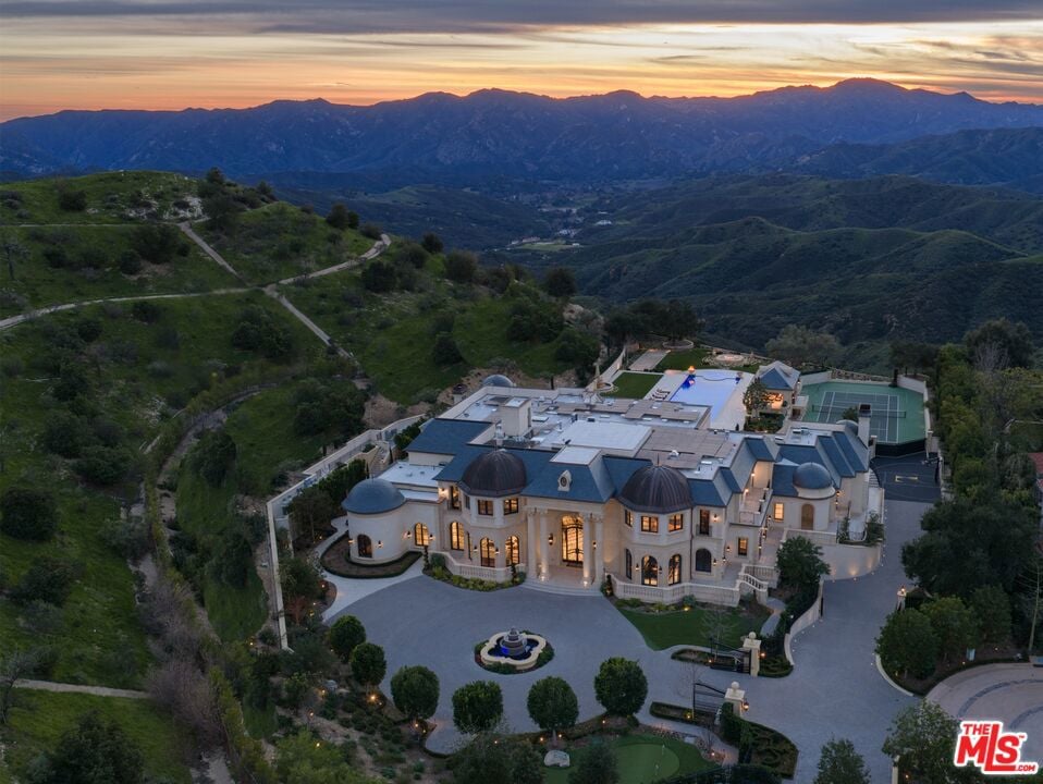 Aerial view of a hillside estate with a pool, tennis court, and mountain sunset.