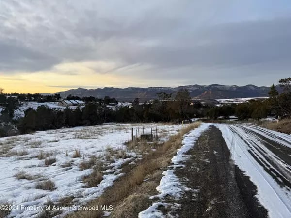 Snowy rural road winds toward a wooded valley and distant mountains beneath a glowing sunset sky.
