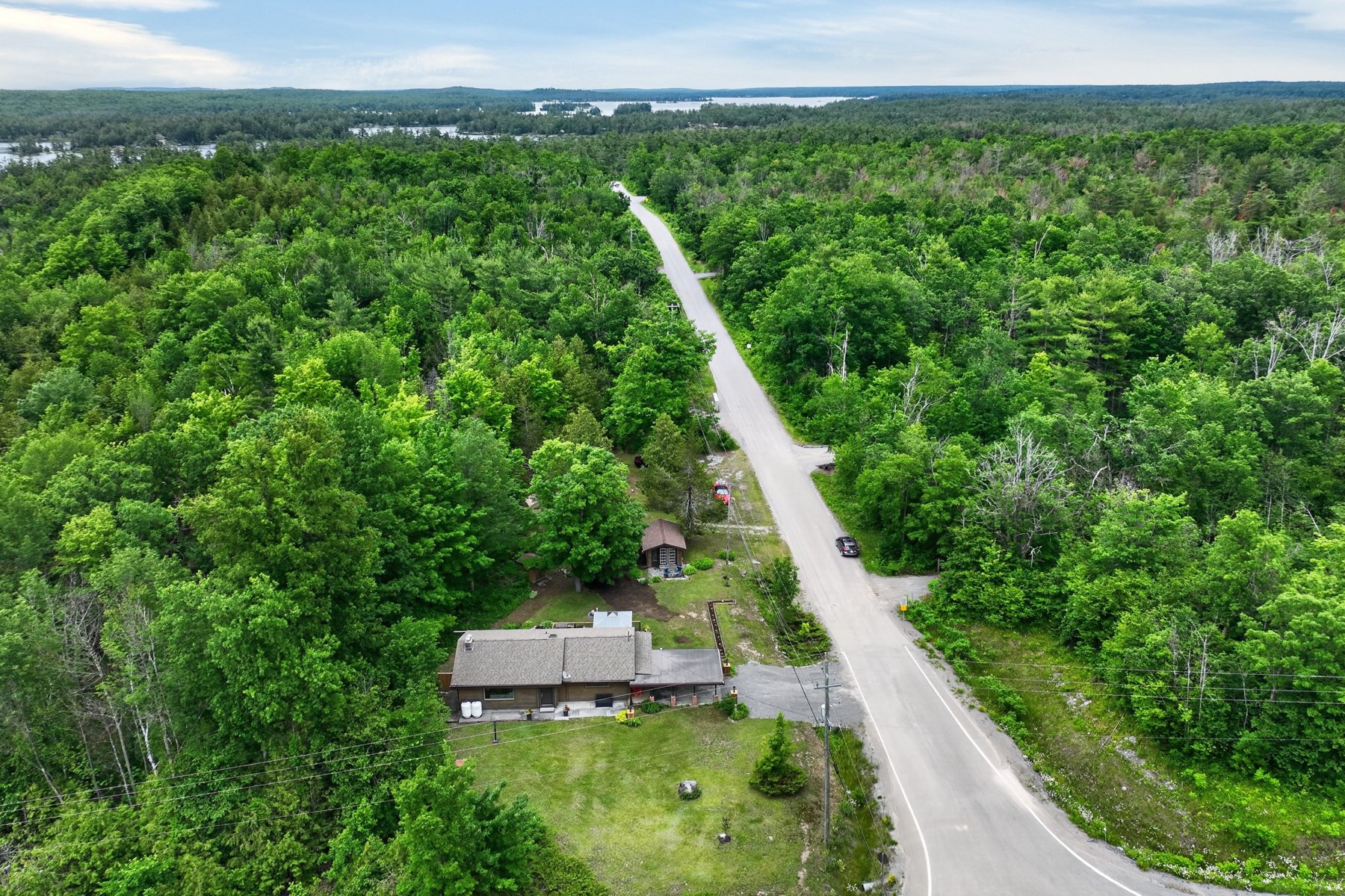 Aerial view of a house beside a rural road, surrounded by dense forest with distant water on the horizon.