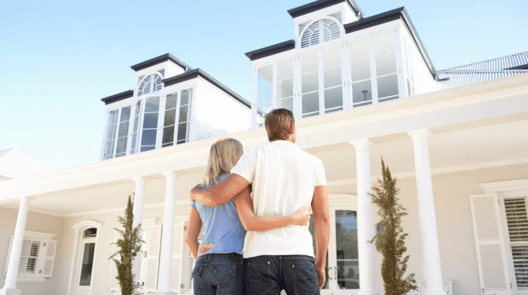 Large white coastal-style house with columns and tall windows, viewed by a couple standing arm in arm.