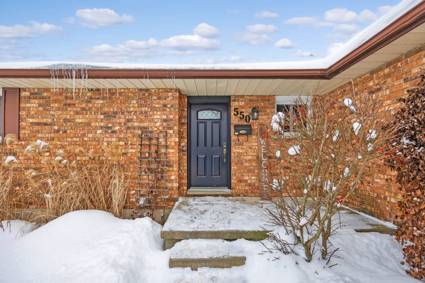 Brick home entrance with a dark blue door, snow-covered steps, and winter landscaping.