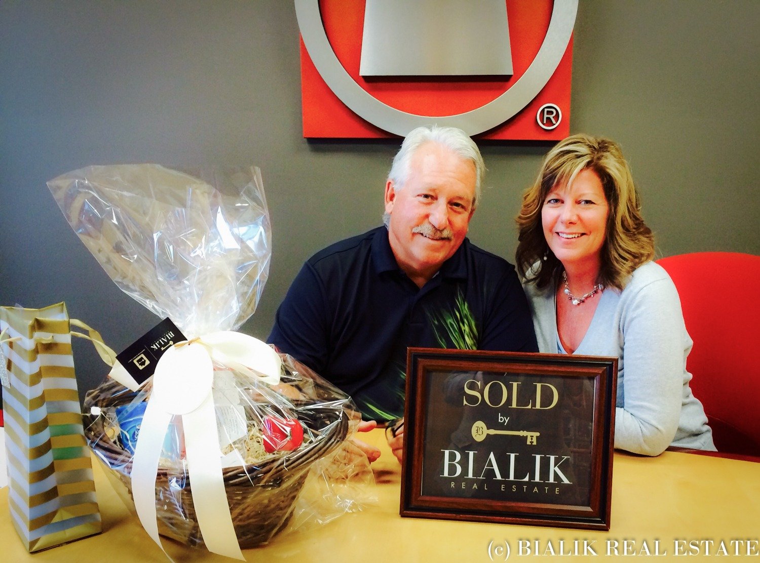 A man and woman celebrate a home sale beside a gift basket and framed sold sign.