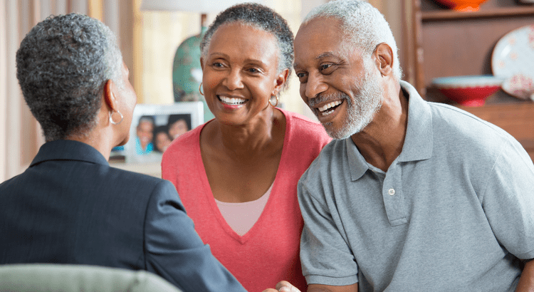 Smiling older couple sits across from a person in a suit, shaking hands in a cozy room. The atmosphere is warm and positive, indicating a friendly meeting.