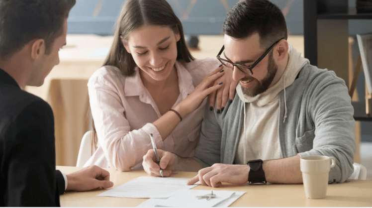 A smiling couple signs paperwork with a real estate agent across the table.