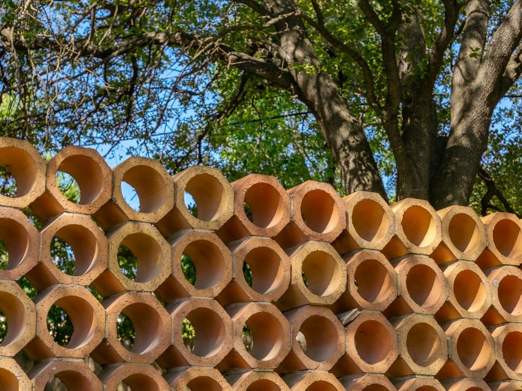 Stacked hollow clay tubes form an outdoor wall beneath leafy trees.