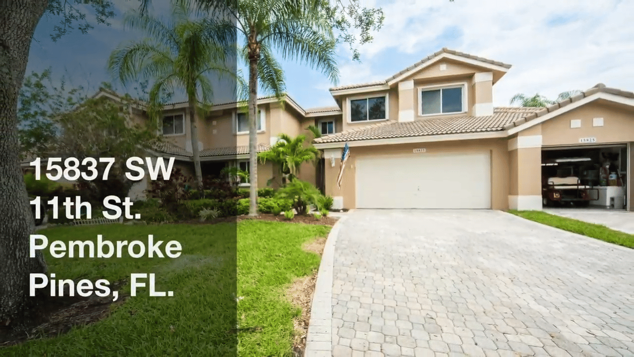 Two-story beige home with a curved paver driveway, palm trees, and an attached garage.