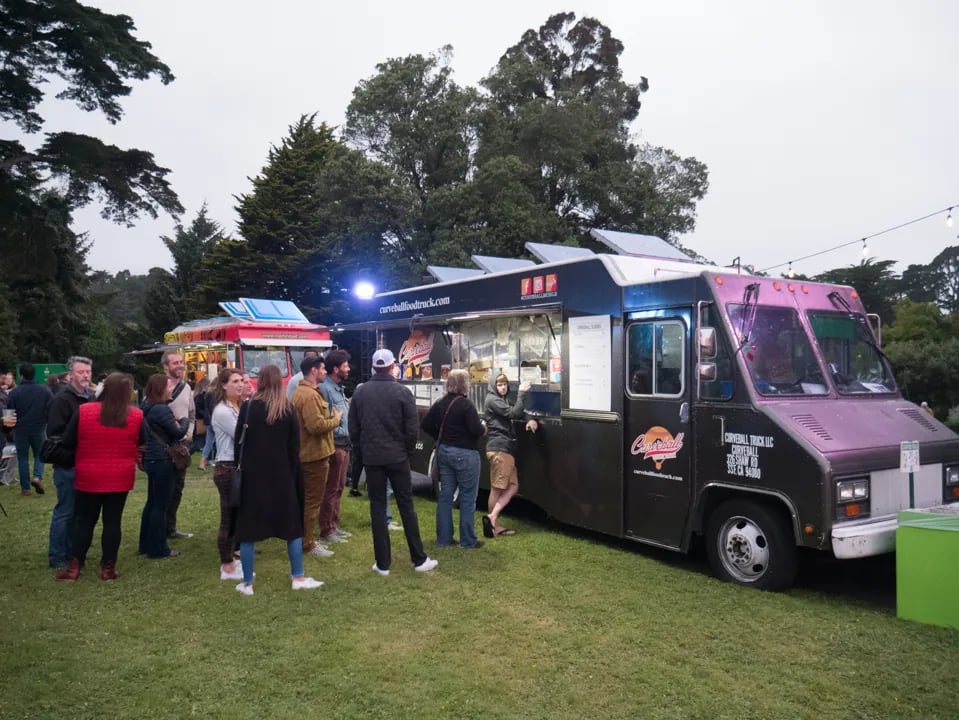 Food trucks lined up on grass with people waiting in line, trees and string lights visible in background