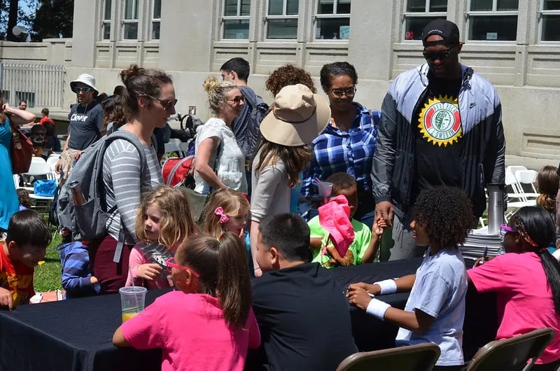 A bright, outdoor scene at the Bay Area Book Festival's youth zone. A group of children and their parents are gathered around a long table covered in a black cloth.