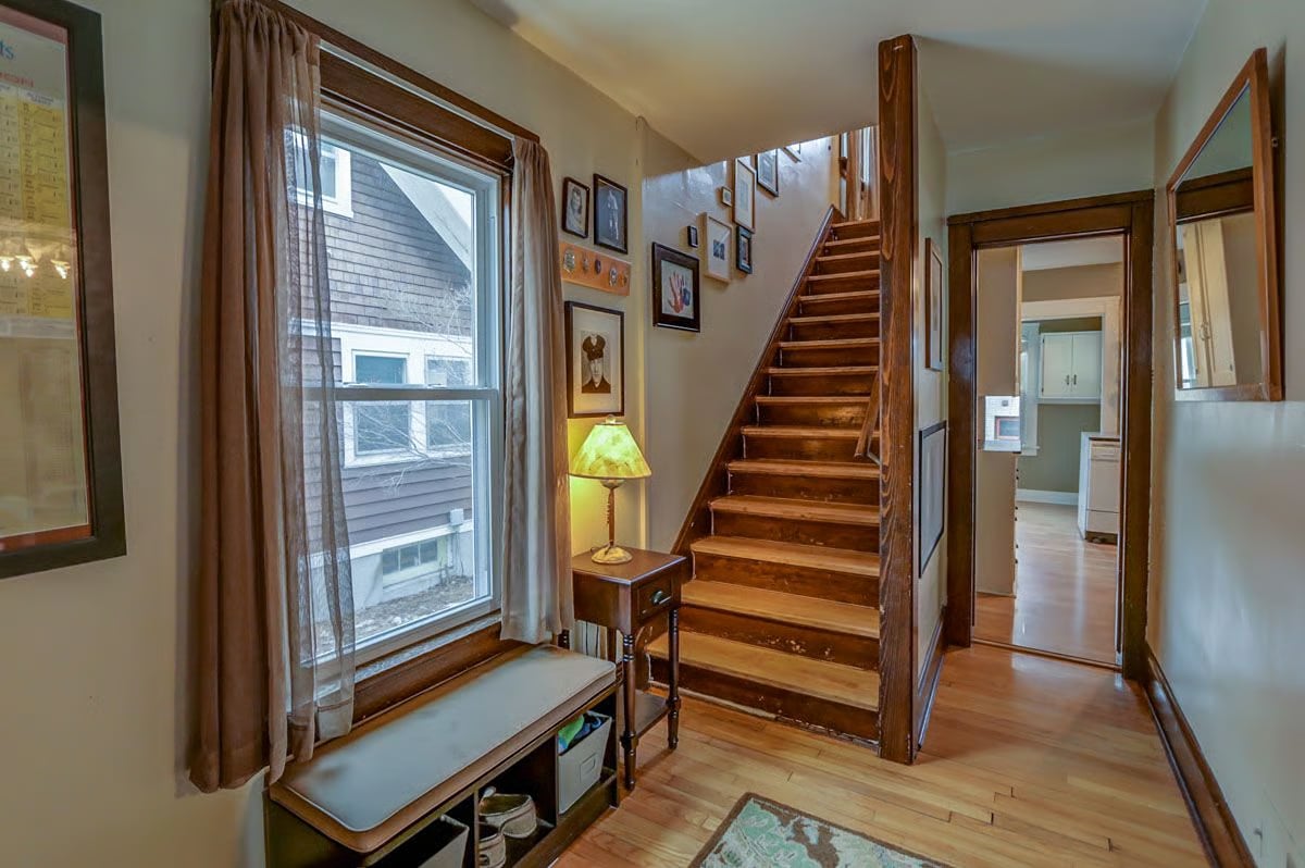 Entry hall with a wide wooden staircase, curtained window seat, and hardwood floors.