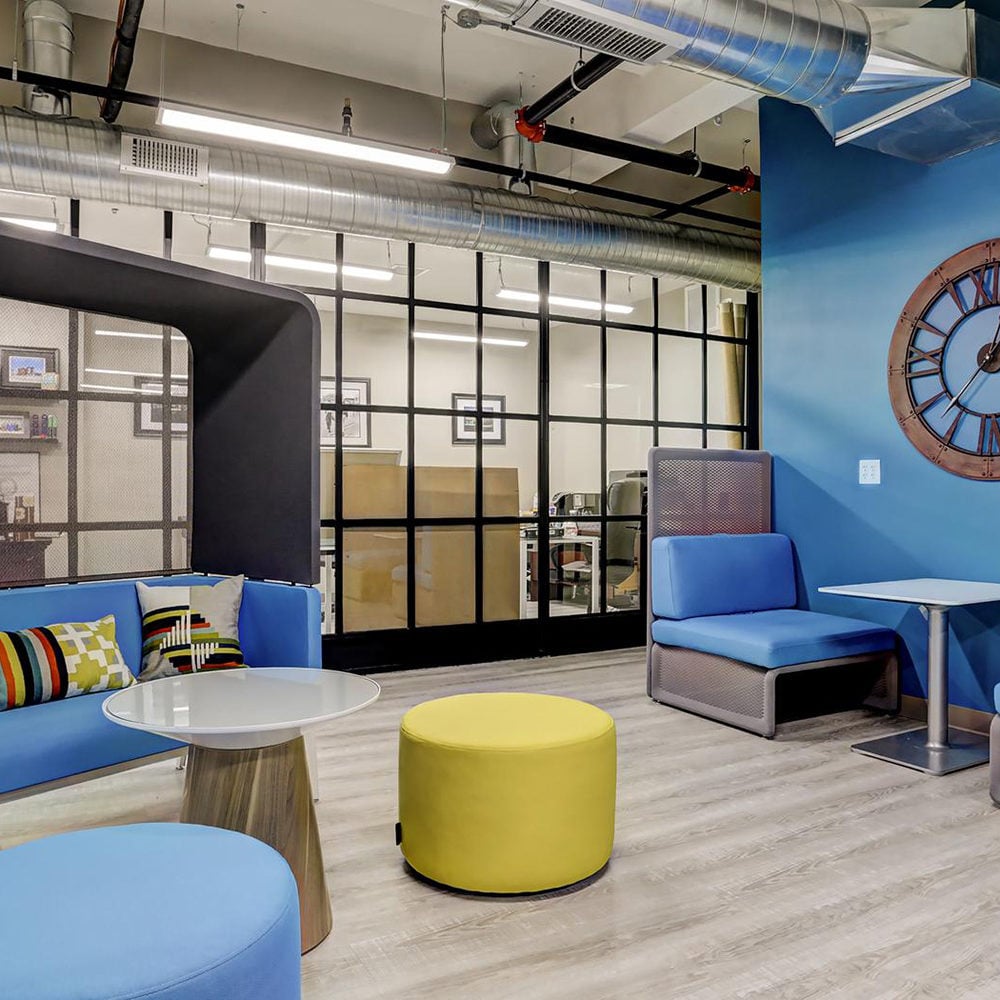 Communal lounge with blue seating, a yellow ottoman, glass partitions, and exposed ductwork.