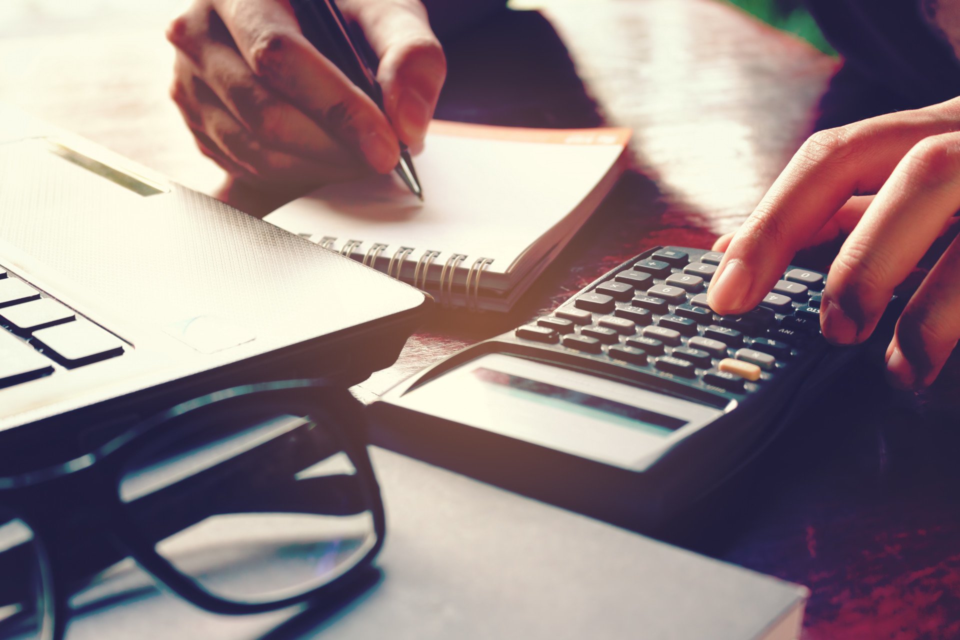 Hands use a calculator and write in a notebook beside a laptop and eyeglasses.