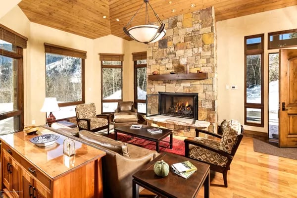 Living room with a stone fireplace, vaulted wood ceiling, leather seating, and snowy views through tall windows.