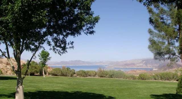 Green park lawn framed by shade trees, with water and distant mountains beyond.