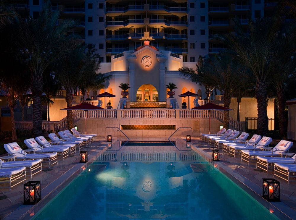 Resort pool at night, lined with lounge chairs and facing the illuminated entrance.