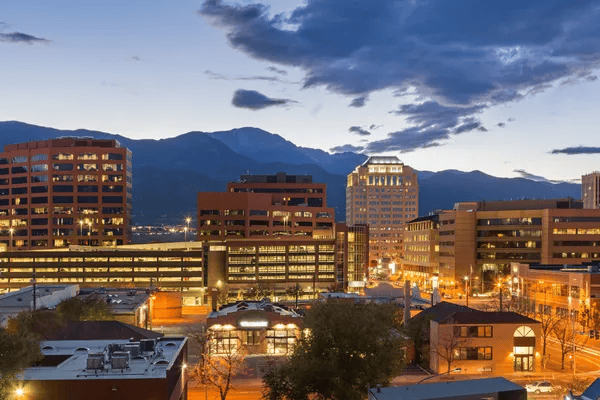 Downtown Colorado Springs glows at dusk beneath the mountains and dramatic clouds.