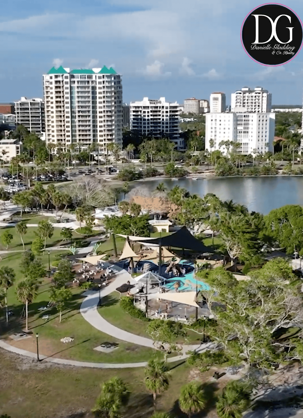 Aerial view of a Sarasota park with a playground and pool beside a lake, backed by high-rise buildings.