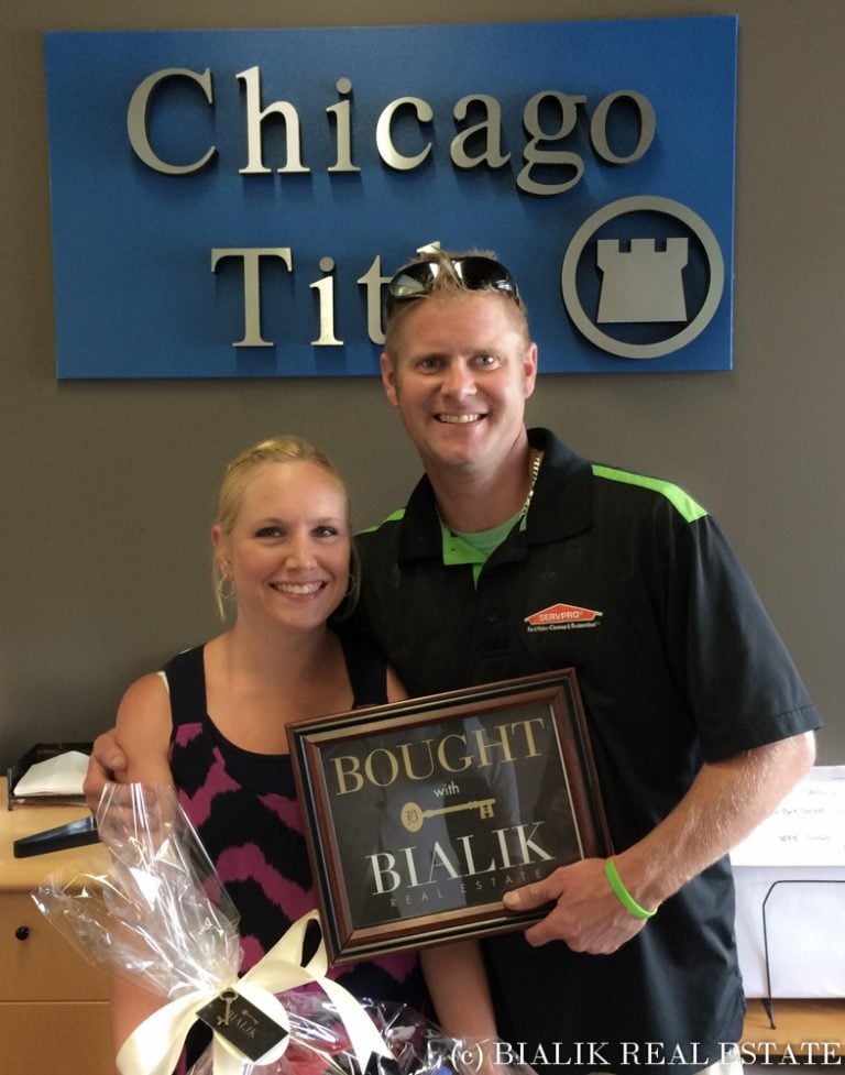 Chad and Erin pose with a framed Bialik Real Estate bought sign and a wrapped gift.