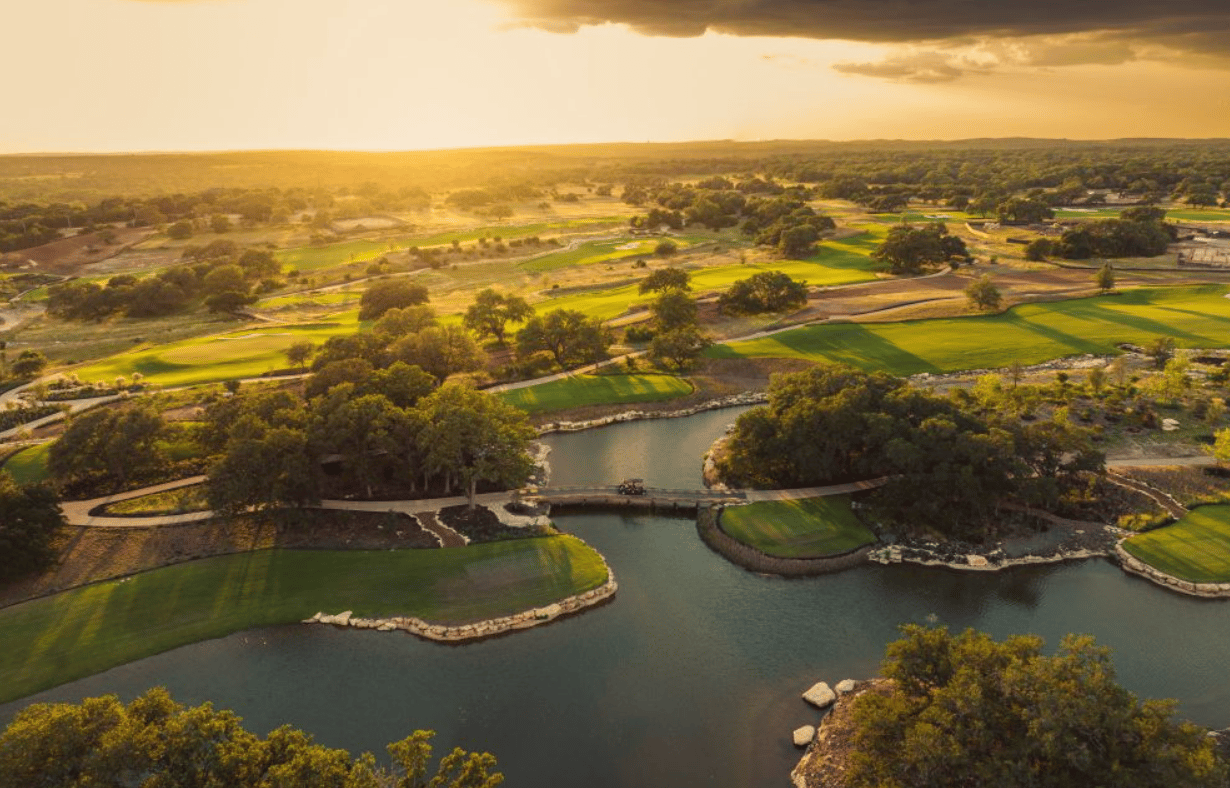 Aerial golf course with a river, landscaped fairways, and wooded countryside in golden-hour light.
