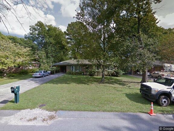 Single-story house behind mature trees, with a broad lawn, driveway, and parked cars.