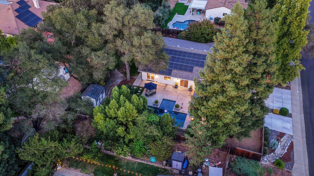 Aerial view of a solar-paneled home with a pool, patio, and mature trees.