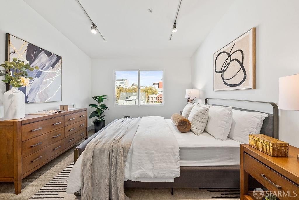 Bright staged bedroom with a white bed, wooden furniture, abstract art, and a window overlooking buildings.