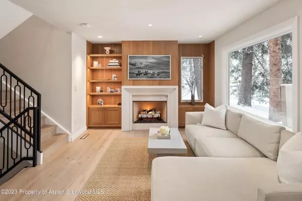 Living room with a white sectional, built-in fireplace and snowy trees visible through large windows.