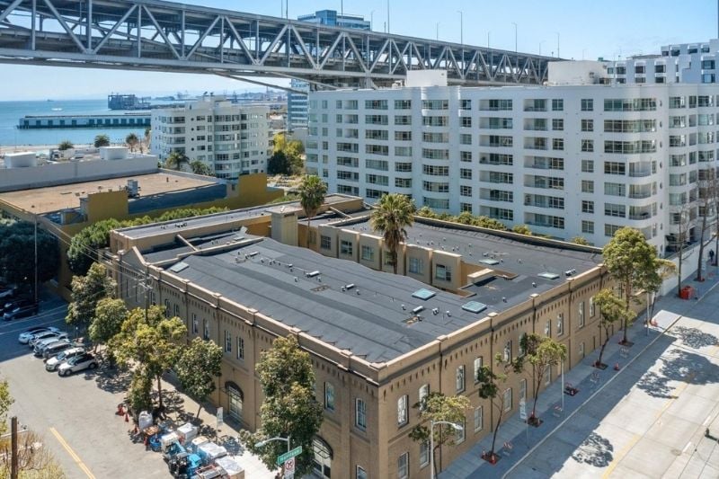 An aerial view of the Harbor Lofts building, showing its brick structure and rooftop, with the San Francisco Bay Bridge and other city buildings in the background.