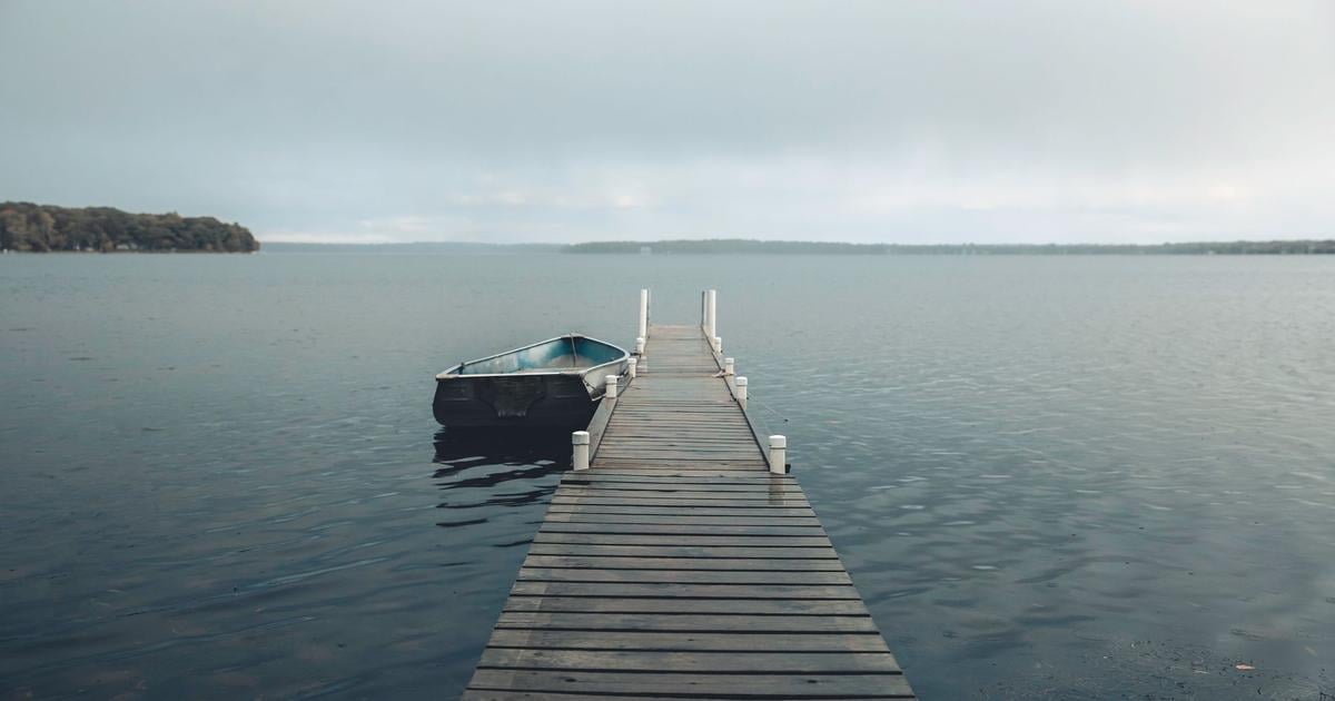 Wooden plank boat dock extending into calm lake water under an overcast sky, a small aluminum boat tied alongside