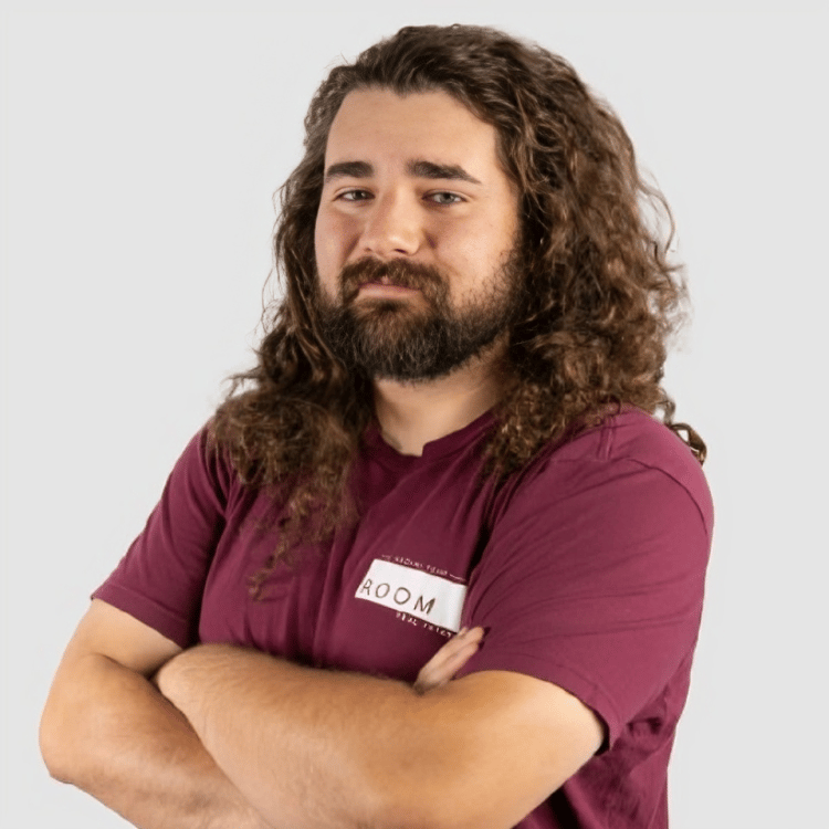 Bailey McDermott with long curly hair and a beard, arms crossed in a maroon ROOM shirt.