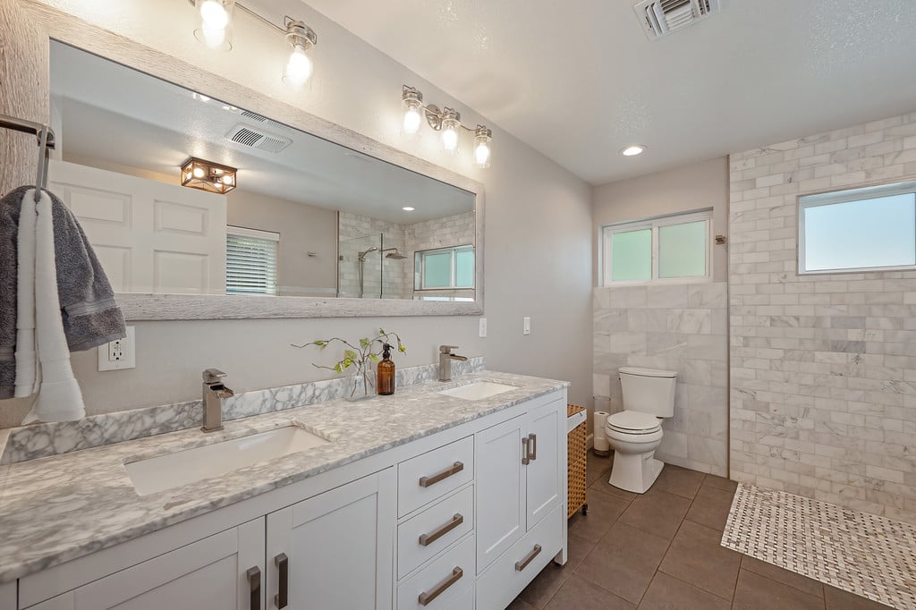 Bathroom with a long white vanity, marble-look counters, tiled shower, and two windows.
